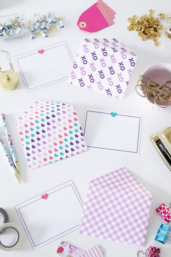 3 white love note cards each one is trimmed with a pink, purple or teal border with a heart at the top in the middle, there are also three DIY envelopes one is covered with xo's, one with various colored hearts at a diagonal, and the final one is a purple gingham print 