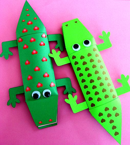 two green pillow box valentine alligators with goggly eyes one has red hearts and the other has black hearts on it