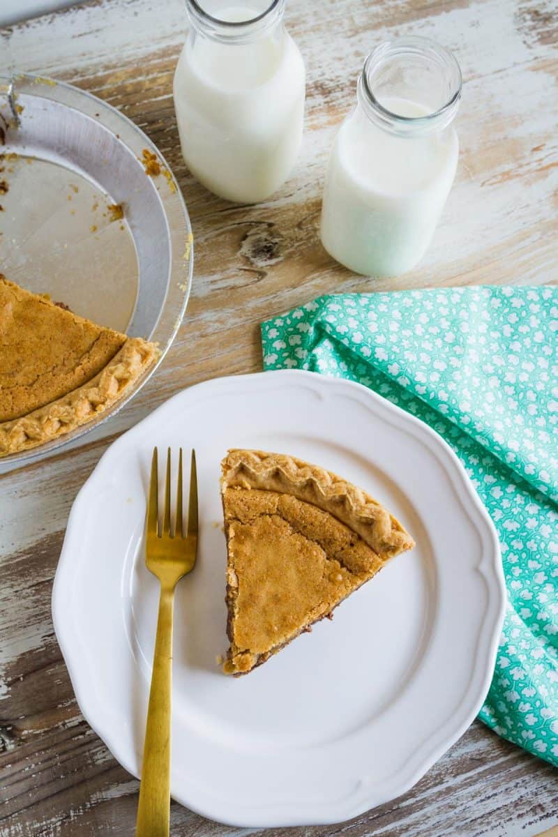 Chocolate Chip Cookie Pie slice on a dish with a gold pie. Jars of milk and half of pie in in the background.  -Skip To My Lou