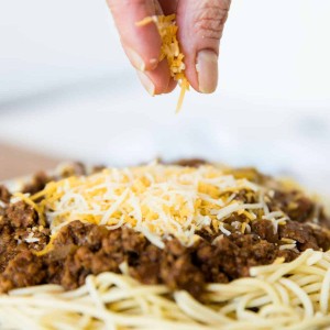 Cincinnati chili served on spaghetti with shredded cheese being sprinkled on to it; served in a white with blue trim detail bowl