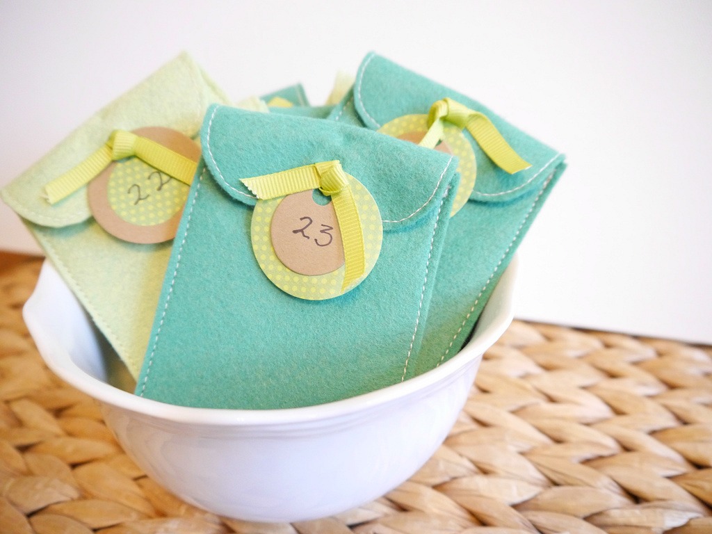 Felt Advent Calendar felt pockets displayed in a white bowl. Pockets have a flap tied with a ribbon and a date tag. -Skip To My Lou