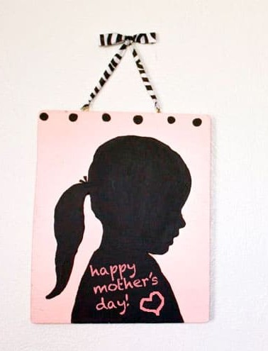 beatiful mother's day gift idea - silouhette chalkboard, the silhouette is of a little girl with a pony tail painted onto a pink background with chalkboard paint with a hanger made out of white and black ribbon
