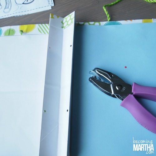 Cut opened gift bag shown with foam board, cardstock, and handle attached. A handheld hole punch is laying on top of the project.