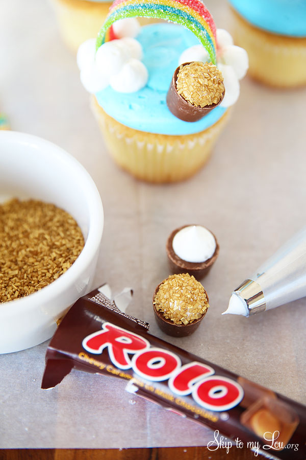 pot of gold made of a Rolo, white frosting and course gold colored sugar for cupcakes; there is a pot of cold added to the top of the cupcake and one decorated beside the cupcake; a package of rolos, piping bag with tip, and bowl of course gold sugar set beside the cupcake