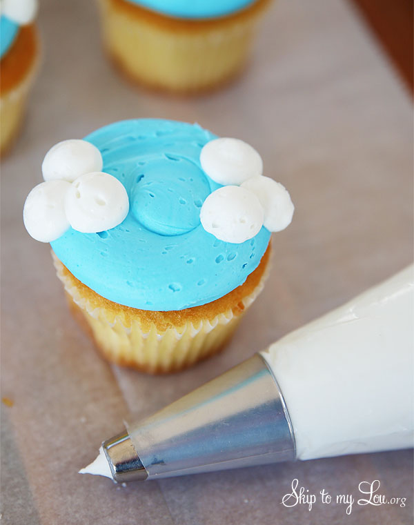 cupcake with sky blue frosting and two sets of three white clouds on two opposite sides of the the cupcake; there is a piping bag with tip beside the cupcake