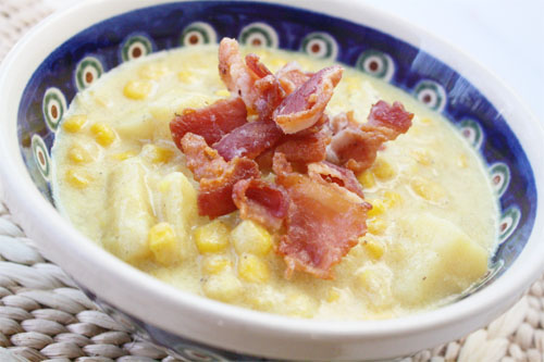 In a beautiful white bowl trimmed in blue with circles of red and green inside white circles is cheddar corn chowder soup garnished with bacon