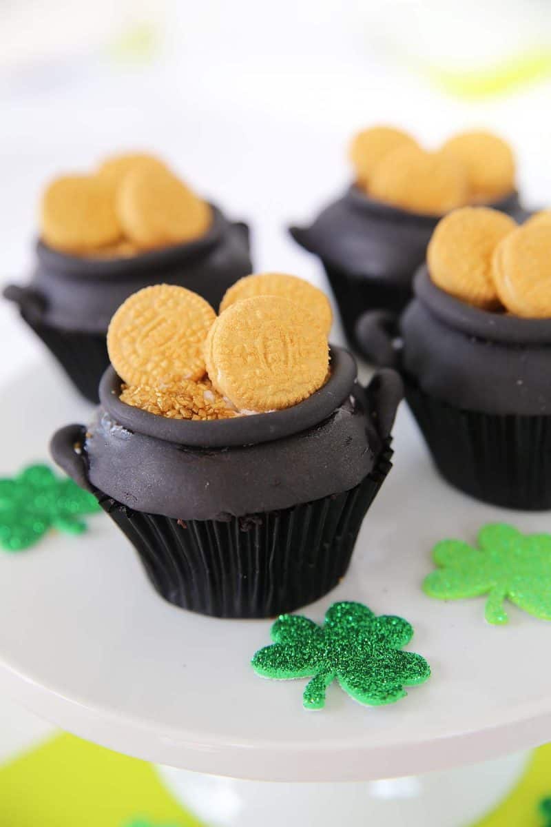 four pot of gold cupcakes with glitter shamrocks lying beside them on a white serving platter