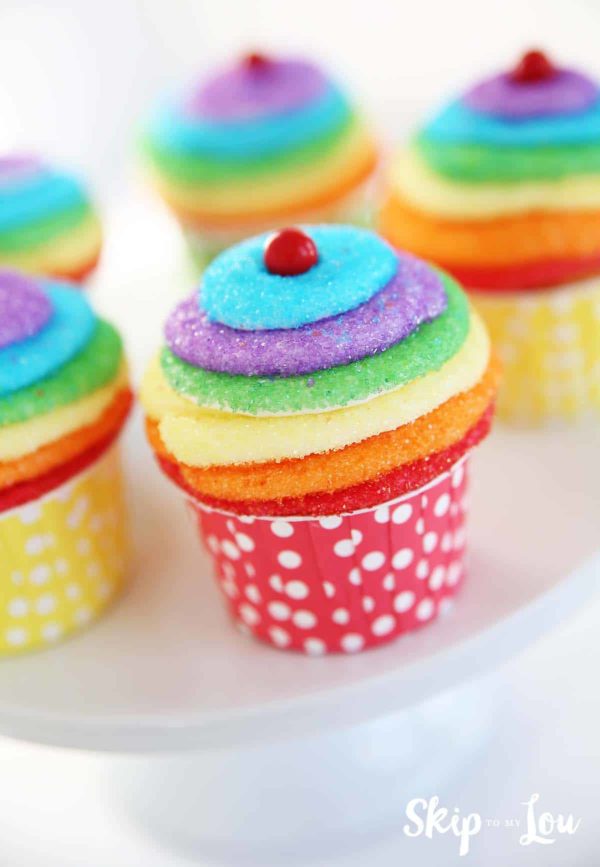 5 cupcakes decorated with rainbow colored layers of frosting; the layers are white frosting, each layer is dipped in the following colors of sanding sugar: red, orange, yellow, green, purple, and blue. topped with  a round red candy in the center