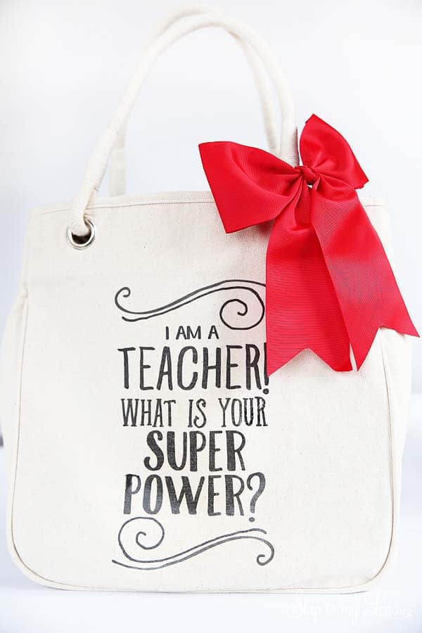 white canvas tote with printable "i am a teacher what is your super power?" transfer sheet applied, there is a red bow attached to the shoulder strap