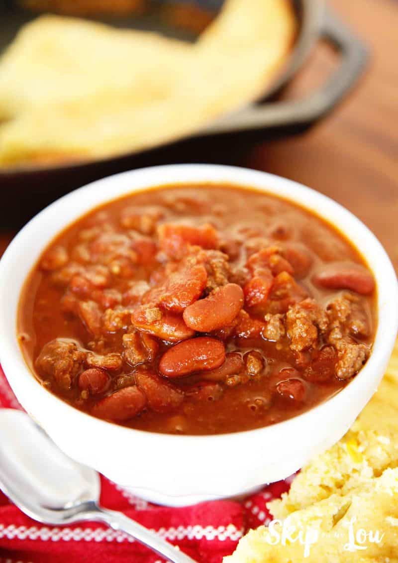 A serving of easy pressure cooker chili in a white bowl with a pan of cornbread in a cast iron skillet in the background. -Skip To My Lou