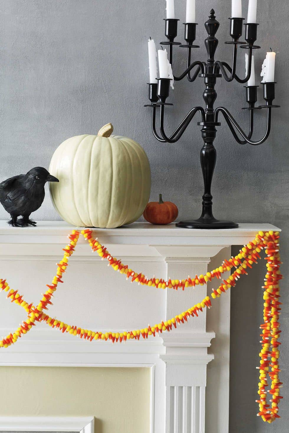 Candy corn garland strewn on a white mantel that is decorated with a black crow, white pumpkin, mini orange pumpkin, and candelabra