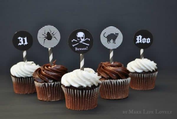 Free printable black and white cupcake picks with the number 31, a spider, skull with word beware printed on it, a black cat, and the word boo on them.  The cupcakes in the image are chocolate with white and brown frosting.