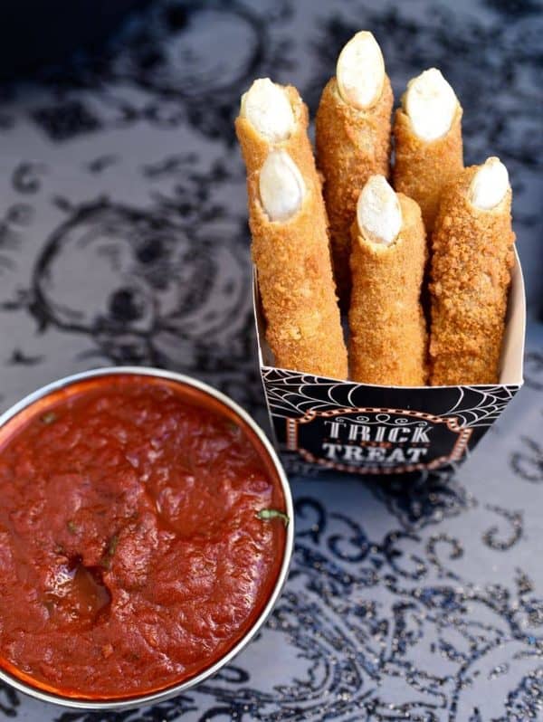 Image shows six mozarella sticks shaped like witch's fingers with a red sauce next to it.