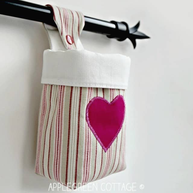 Pink and beige striped fabric hanging storage basket with fuschia pink heart sewn on the front and a button strap to hang it with; the basket has white trim and lining; it is hung on a black rod