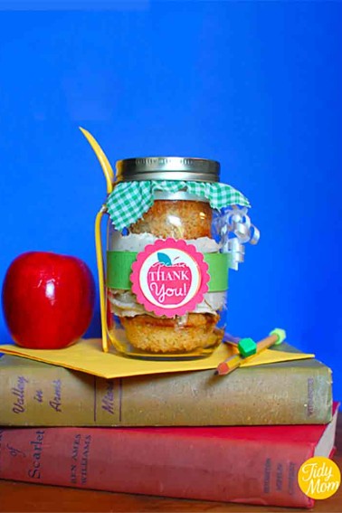 cupcakes in a jar with an apple thank you free printable tag wrapped the jar, the lid has a piece green gingham printed fabric over the top and a plastic fork is attached to the jar; this is reting on two books, and a card with a red apple to the left and pencil to the right