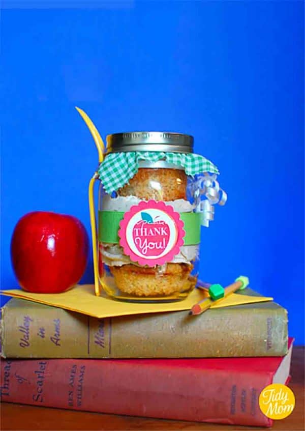 cupcakes in a jar with an apple thank you free printable tag wrapped the jar, the lid has a piece green gingham printed fabric over the top and a plastic fork is attached to the jar; this is reting on two books, and a card with a red apple to the left and pencil to the right