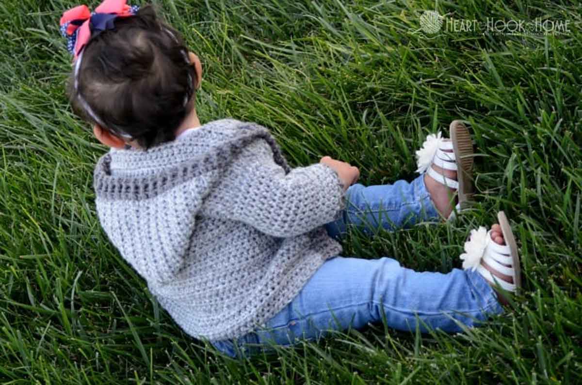 Image shows a baby sitting in the grass wearing a hoodie crochet grey sweater. -Skip To My Lou