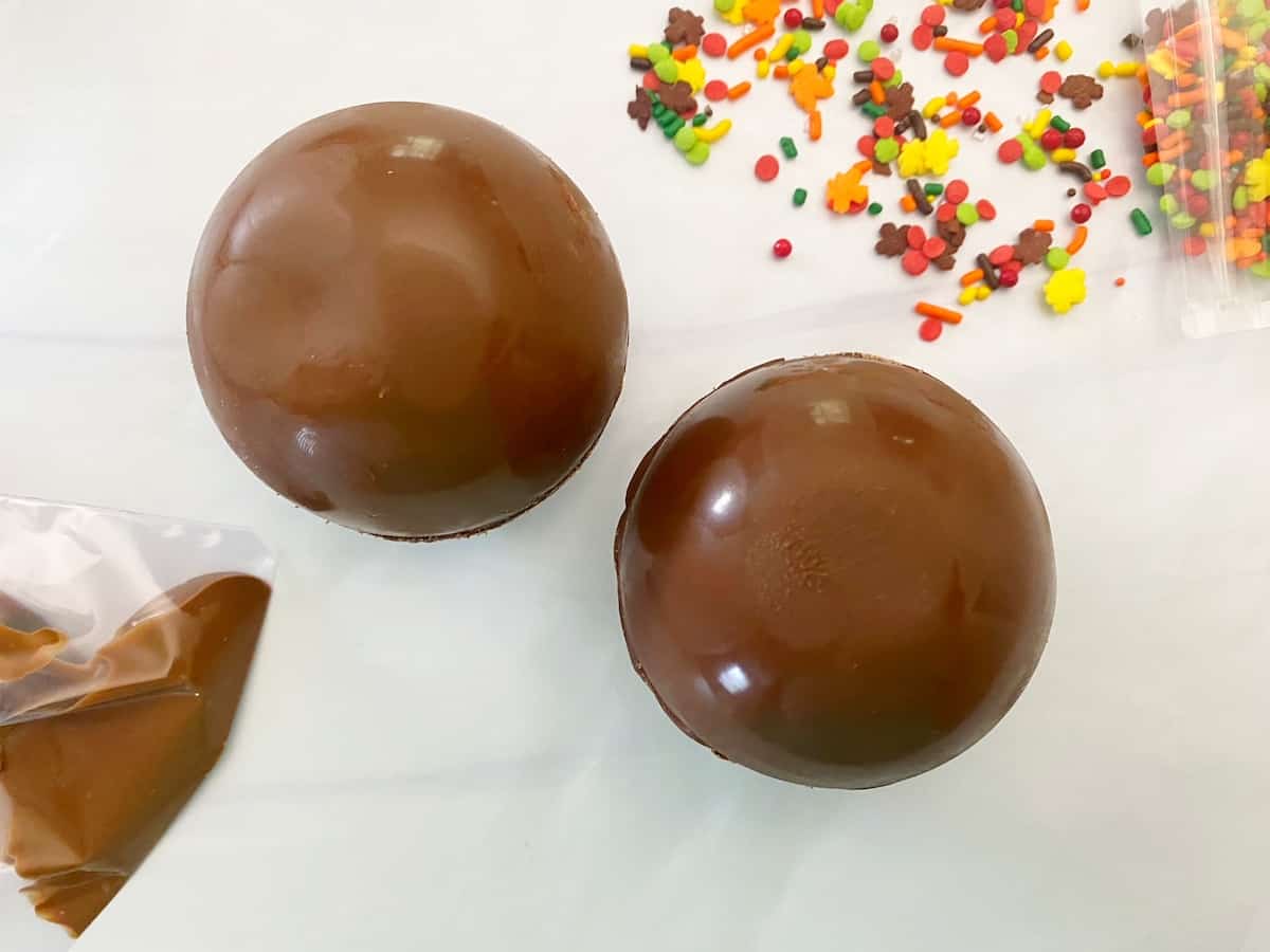 chocolate cocoa bombs with sprinkles to side and pastry bag with melted chocolate to side
