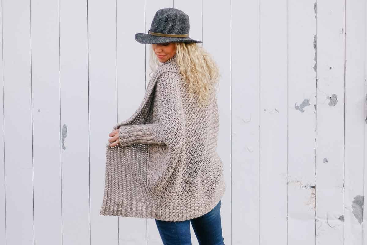 Image shows a blonde woman wearing a DIY crochet sweater standing in front of a wooden wall. -Skip To My Lou