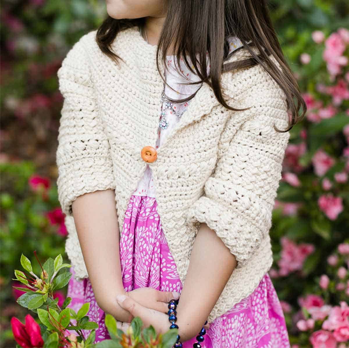 Image shows a little girl wearing crochet cardigan in front of a blurry background. Skip To My Lou