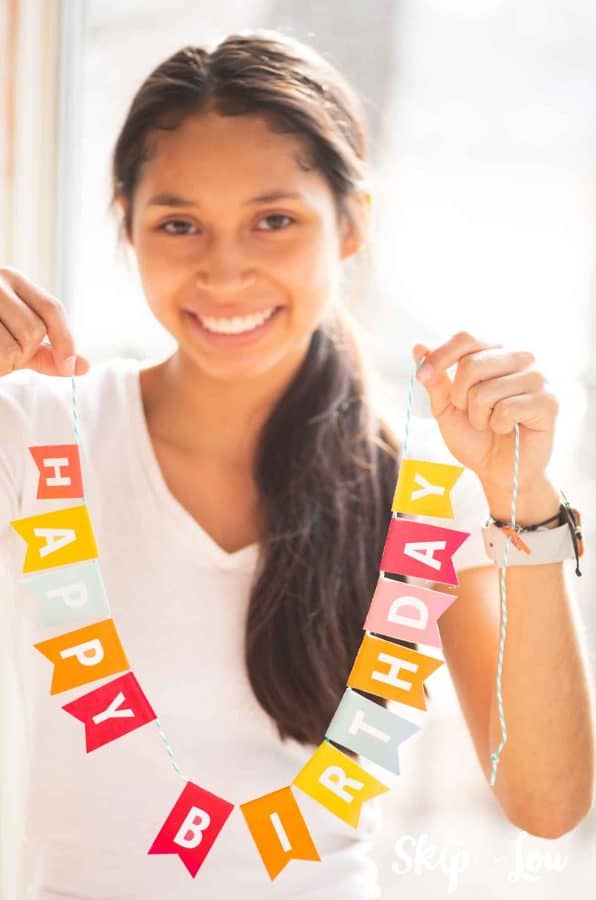 Beautiful young lady smiling and holding a colorful banner that says Happy Birthday by Skip to my Lou.
