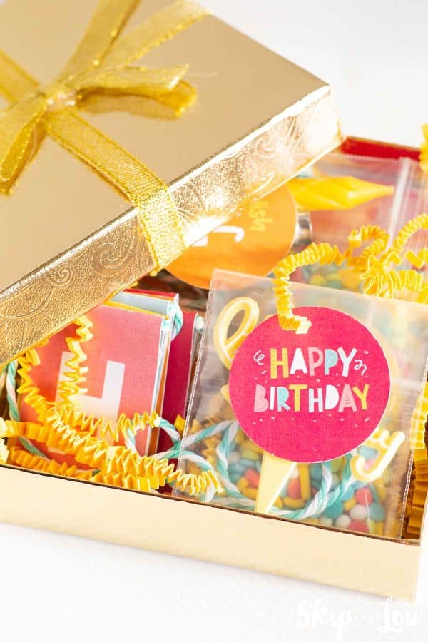 Gold foil box with the lid half off revealing the contents of the tiny birthday box. Contents are a Happy Birthday banner, sprinkles, a candle and lighter and colorful paper by Skip to my Lou.