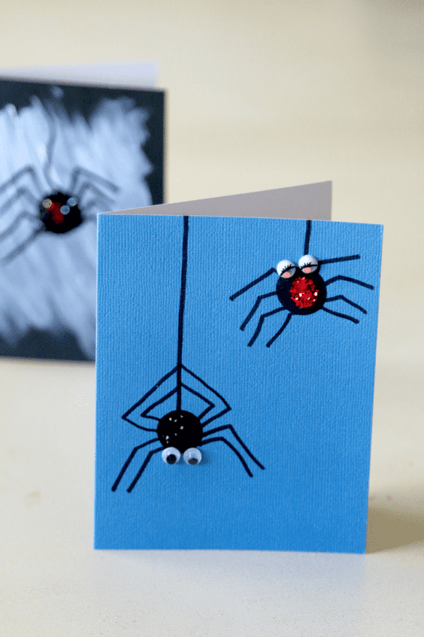 Two black thumbprint spiders with googly eyes hanging from a spider web on a blue piece of folded cardstock paper.