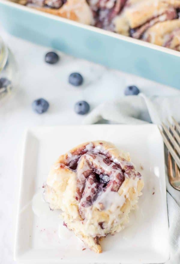 Image shows a sweet cinnamon roll served on a plate with single blueberries next to it.