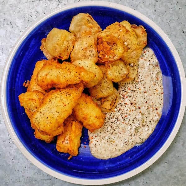 Iage shows a sourdough tempura fried pacific cod with dressing on a blue plate.