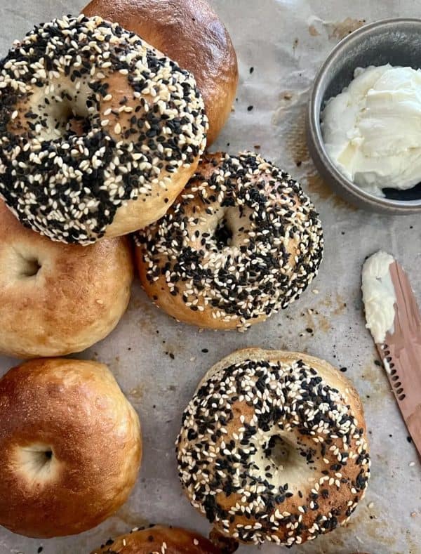 Image shows three sesame seed sourdough bagels on a table with a wooden knife next to them.