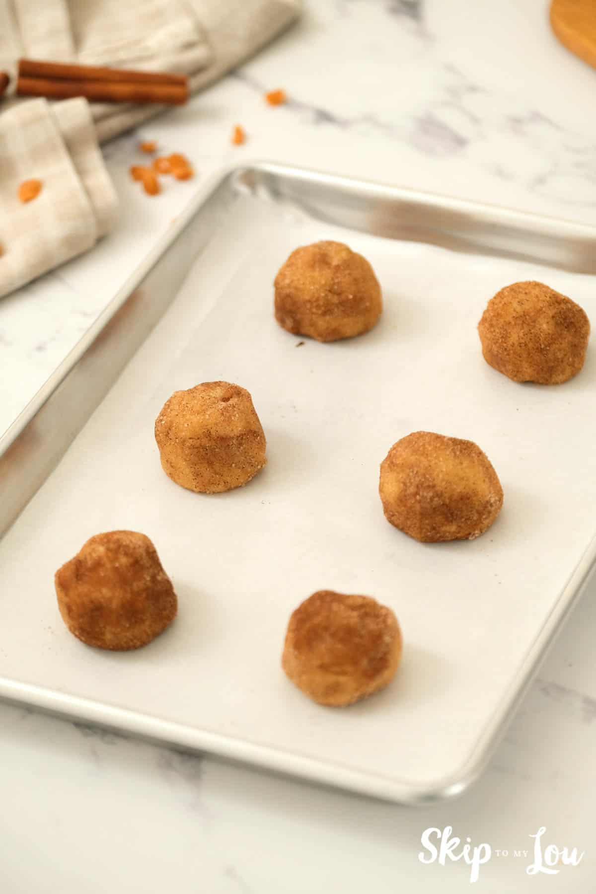 6 balls of cookie dough covered in a cinnamon sugar mixture on a parchment paper covered baking sheet. Toffee Doodle Cookie Recipe -Skip To My Lou