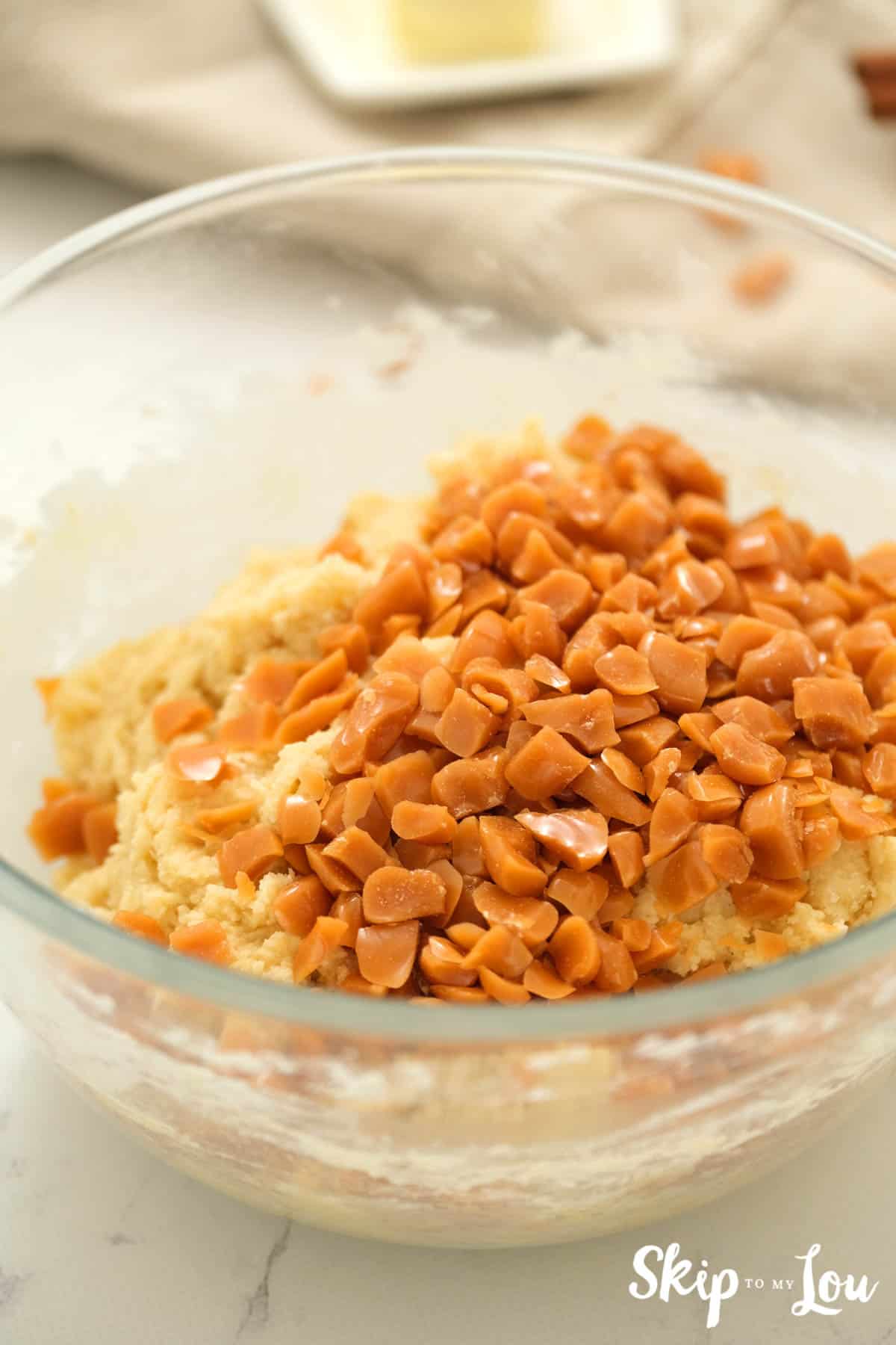 Large clear glass mixing bowl with batter for Toffee Doodle Cookies with the toffee bits on top of the batter ready to stir in. -Skip To My Lou