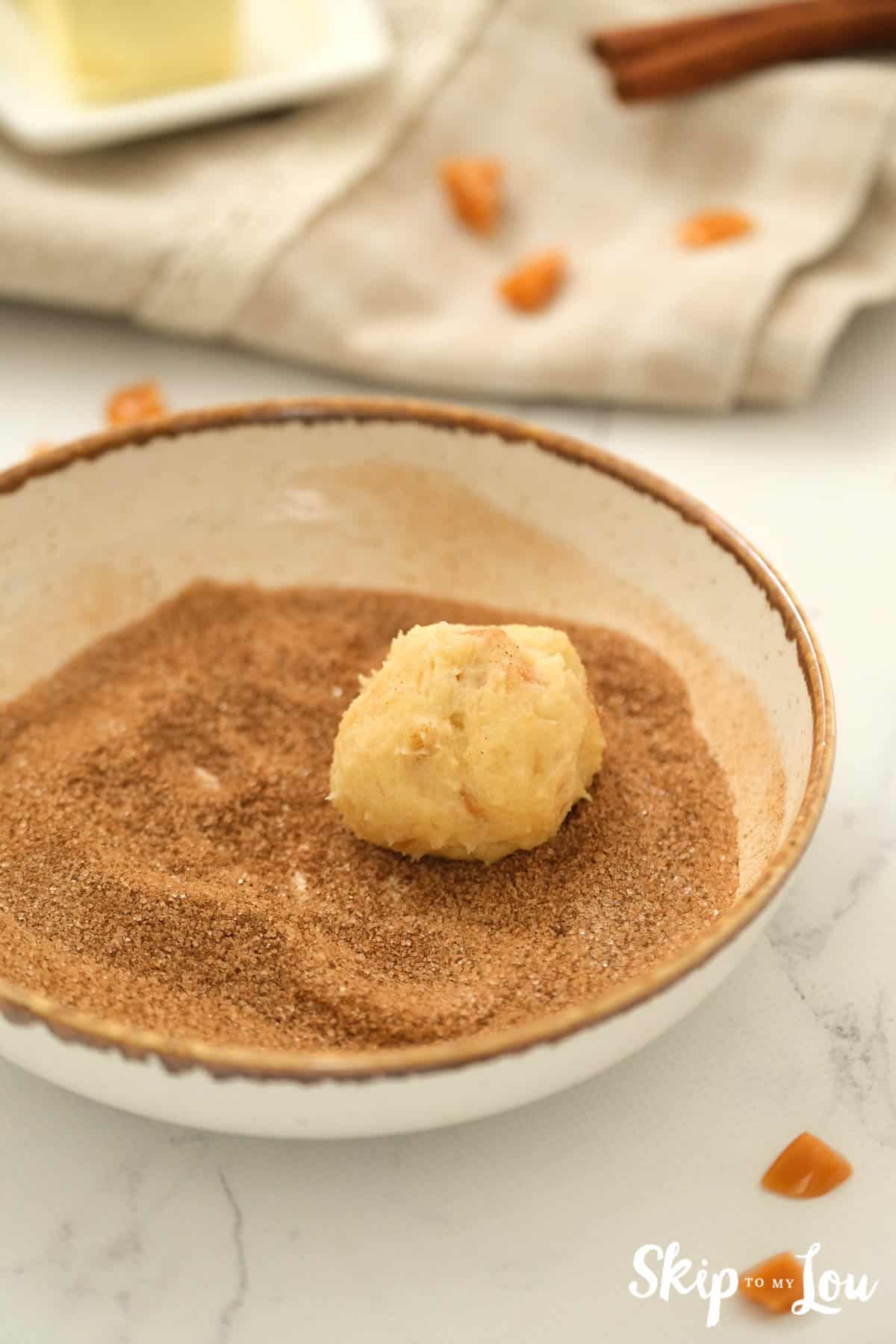 White bowl with sugar cinnamon mixture and ball of cookie dough ready to be rolled in the mixture. Toffee Doodle Cookie Recipe -Skip To My Lou