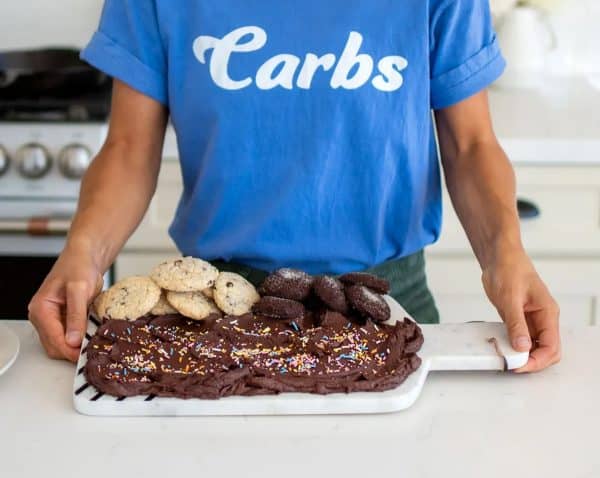 image shows a woman holding a chocolate buttercream board with cookie and muffins inside a kitchen.