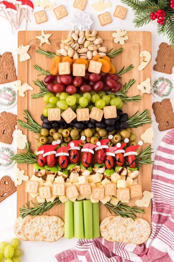 Image shows a chrcuterie board with crackers, vegetables decorated with Santa hats, and cookies that spell out Christmas. via Happy money Saver.
