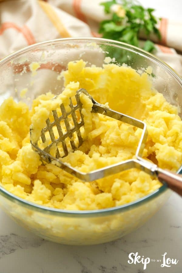 Potatoes that have been mashed in a clear mixing bowl with the masher laid on top. Instant Pot Mashed Potatoes -Skip To My Lou
