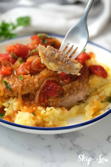 A bite of swiss steak on a fork. The swiss steak recipe is served in a white bowl with a blue rim.