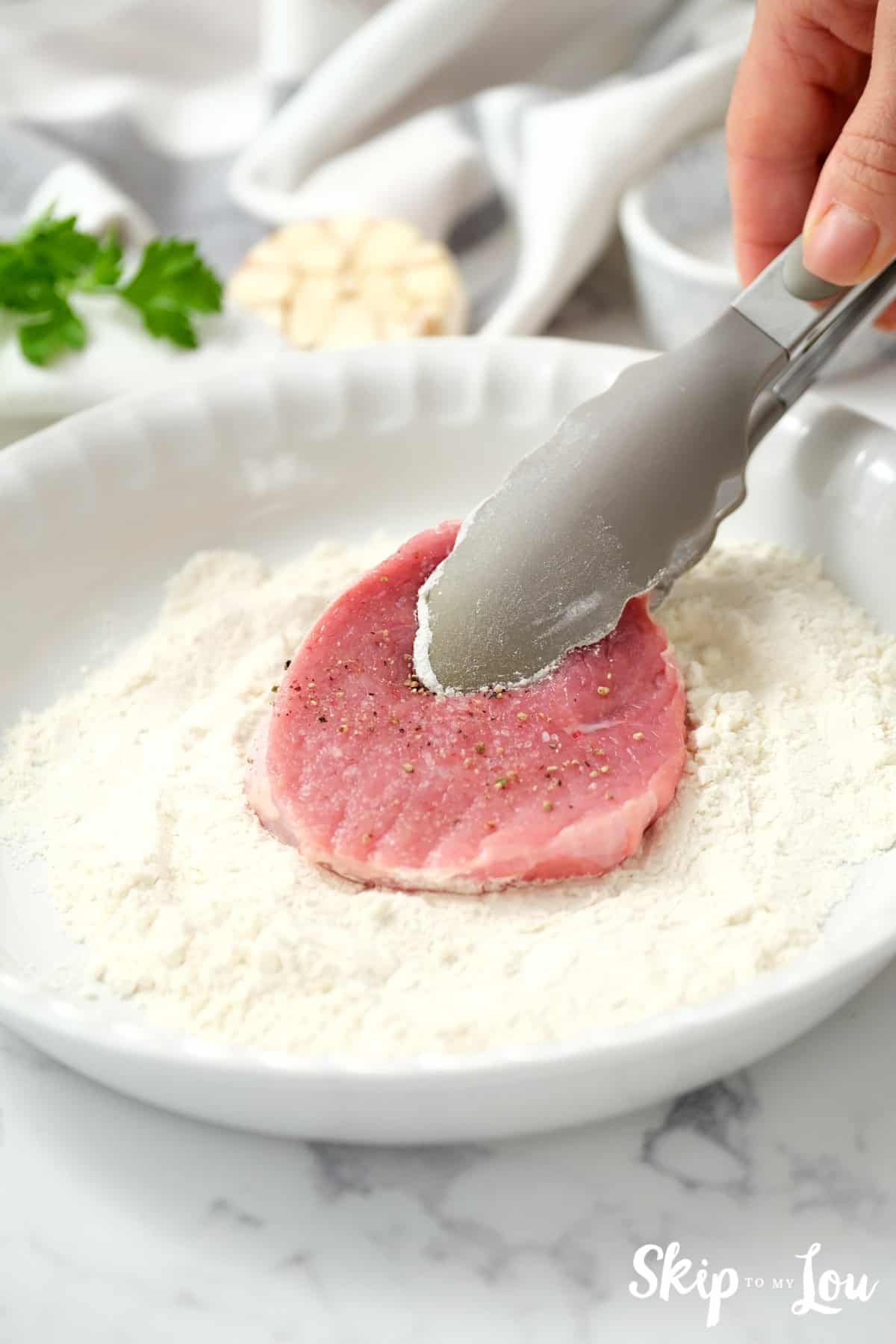 Bottom Round Steak slice being dredged in flour in a white pie pan. Swiss Steak Recipe -Skip To My Lou