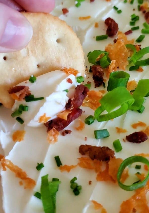 Image shows a cream cheese board with baked potato ingredients and a person's hand eating from it.