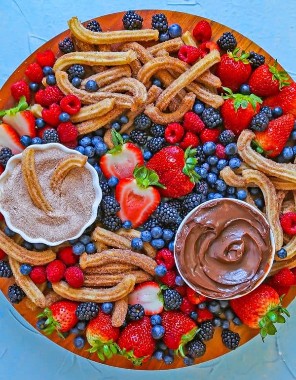 Image shows a large dessert charcuterie board with churros, fruits and chocolate dip. via the comfort of cooking