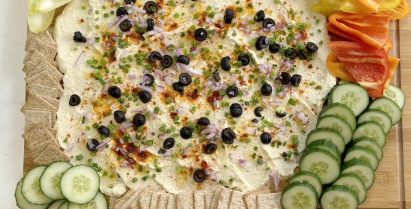 Image shows a mediterranean hummus board with cucumber, red pepper, and black olives.