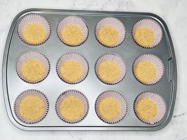 graham cracker crumbs in the bottom of cupcake liners in a muffin pan
