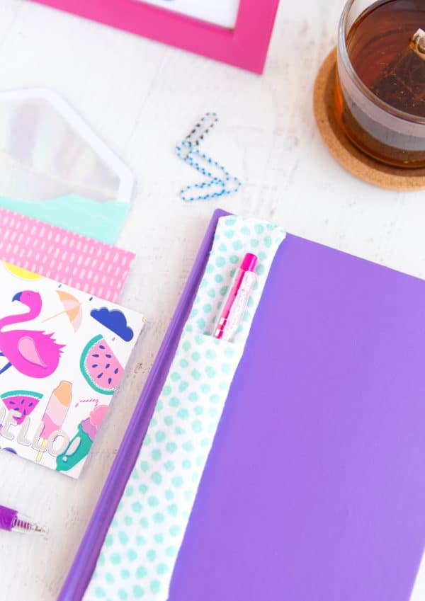 Pen holder with a pink pen inside and on top of a purple notebook and other school supplies.