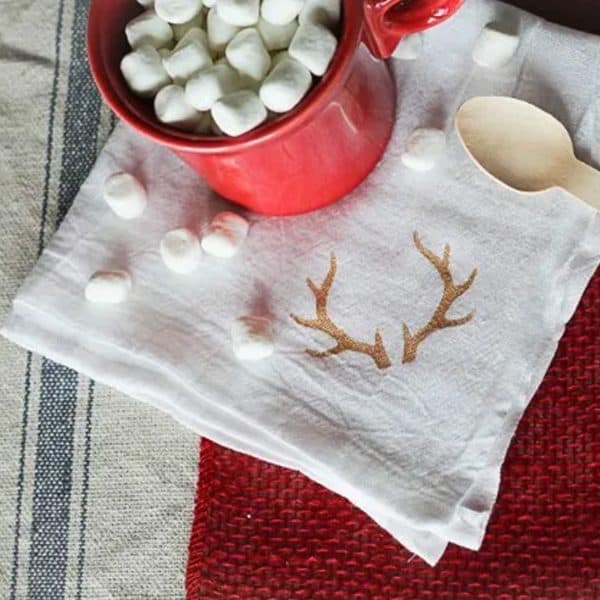 Image shows a DIY antler napkin with a mug with hot cocoa and marshmallows inside.