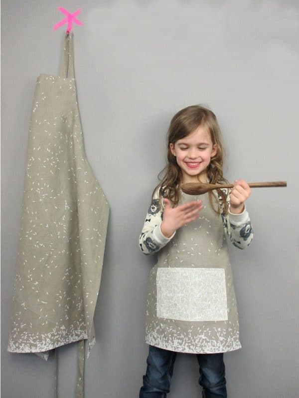 Kid wearing an apron holding a wooden spoon and a larger apron hanging from the wall.