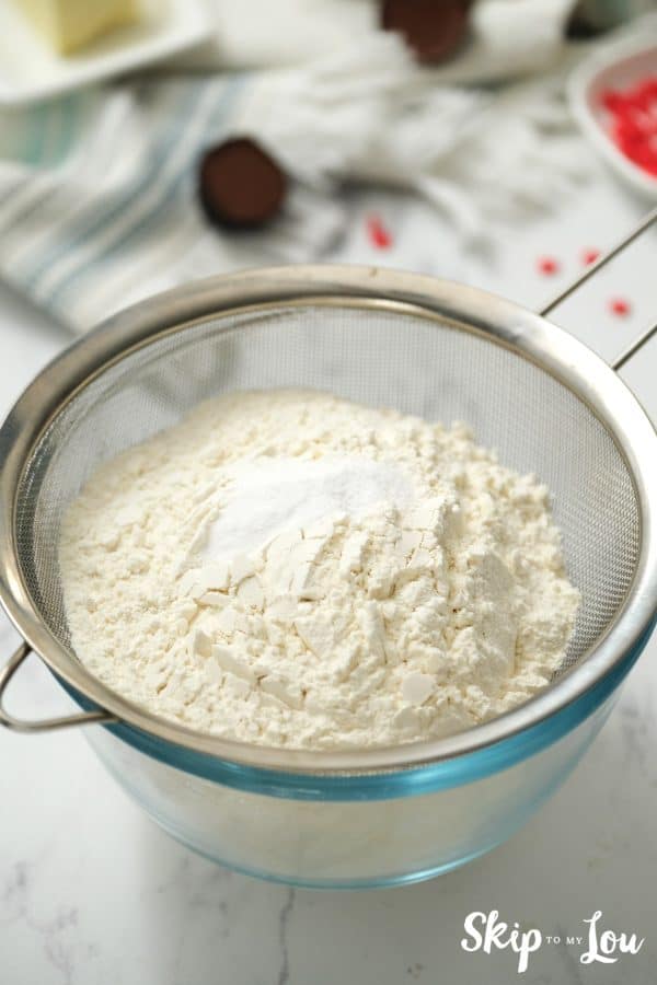 Silver metal sifter over a round glass mixing bowl holding flour, salt, baking soda and sugar, by Skip to my Lou.