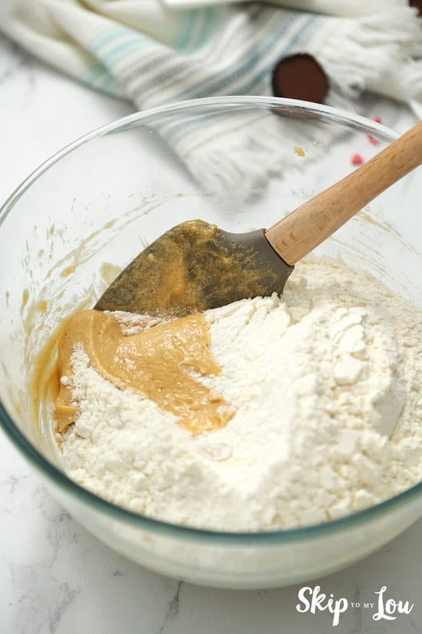 Round glass mixing bowl with flour and peanut butter being mixed by a black spatula, by Skip to my Lou.