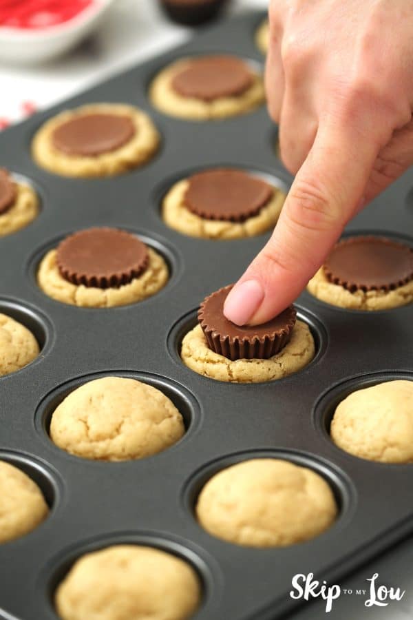 Finger pressing a peanut butter cup candy into the center of a cookie baked in a mini cupcake tin, by Skip to my Lou.
