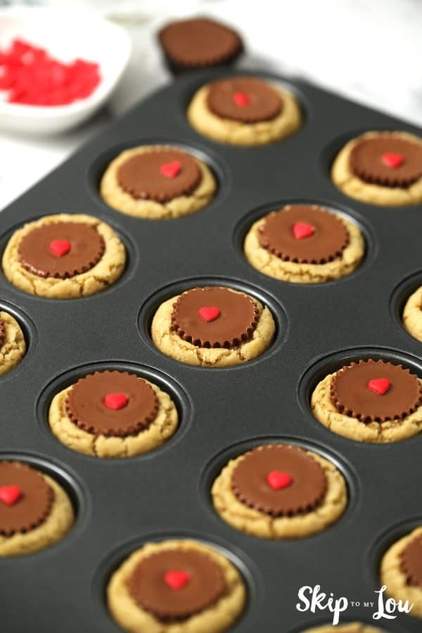 Black mini cupcake tin filled with Reece's Peanut Butter Cup cookies, each having 1 red candy heart in the center, by Skip to my Lou.