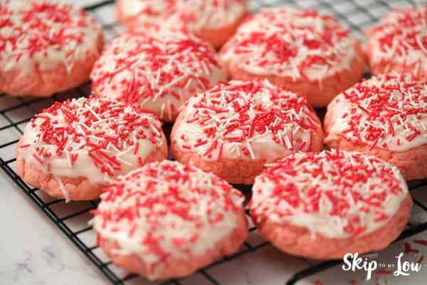 9 strawberry pink cookies with white icing and sprinkles, sitting on a black wire cooling rack, by Skip to my Lou.